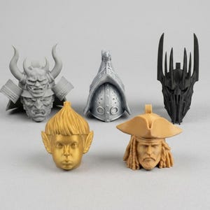 May include: A collection of five miniature sculpted heads and helmets including a grey samurai mask, a silver gladiator helmet, a black spiked crown, a gold-coloured elf head with pointed ears, and a tan pirate head wearing a tricorn hat.