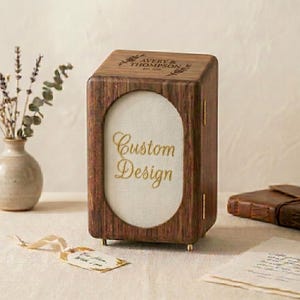 Könnte beinhalten: Eine rechteckige Box aus dunklem Holz mit einem vertikalen ovalen Ausschnitt auf der Vorderseite, die einen cremefarbenen Einsatz mit dem goldenen Text Custom Design enthält. Die Oberseite ist mit AVERY & THOMPSON EST. 2020 und Blumen graviert.