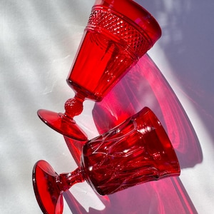 May include: Two red vintage-style wine glasses with intricate patterns. One glass is upright, showcasing a textured rim, while the other lies on its side. The glasses cast deep red shadows on a white surface, highlighting their ruby color.