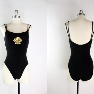 May include: Black one-piece swimsuit with a gold seashell design on the front. The swimsuit has thin, braided straps and a scoop back. The garment is displayed on a mannequin.