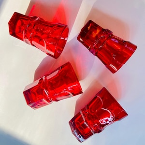 May include: Four red glass tumblers with a textured, geometric design. The glasses are arranged in a semi-circle on a white surface.