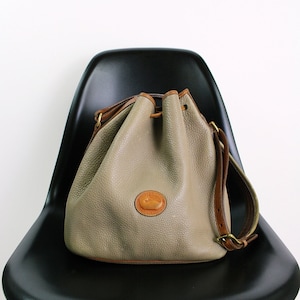 Puede incluir: Un bolso de cuero de color gris claro con una correa de cuero marrón y un logotipo de cuero marrón. El bolso está sentado en una silla negra.