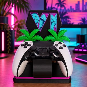 May include: A black and white video game controller resting on a black stand. The stand features a hexagonal backplate with the letters VI in a cyan palm tree pattern, flanked by two bright green plastic palm trees and a neon pink base accent.