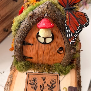 May include: A whimsical fairy house with a wooden door, a red mushroom-shaped light, and a monarch butterfly. The house is decorated with moss, flowers, and a small sign with floral illustrations. A miniature witch hat rests on a book.