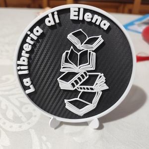 Può includere: Un cartello rotondo con la scritta "La libreria di Elena" e una pila di libri aperti. Il cartello ha un centro nero strutturato e due piedini bianchi per stare in piedi.