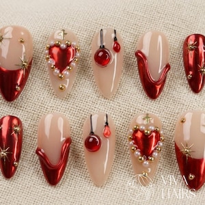 May include: A selection of artificial nails with diverse designs. Some nails have a nude base with red metallic tips, while others display red heart and pearl embellishments. Gold star and bead accents add detail.