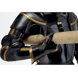 May include: Close-up of a black and gold medieval suit of armor. The armor features intricate gold detailing along the edges and joints. The gauntlets are holding a wooden object. The armor is highly polished and reflects the light.