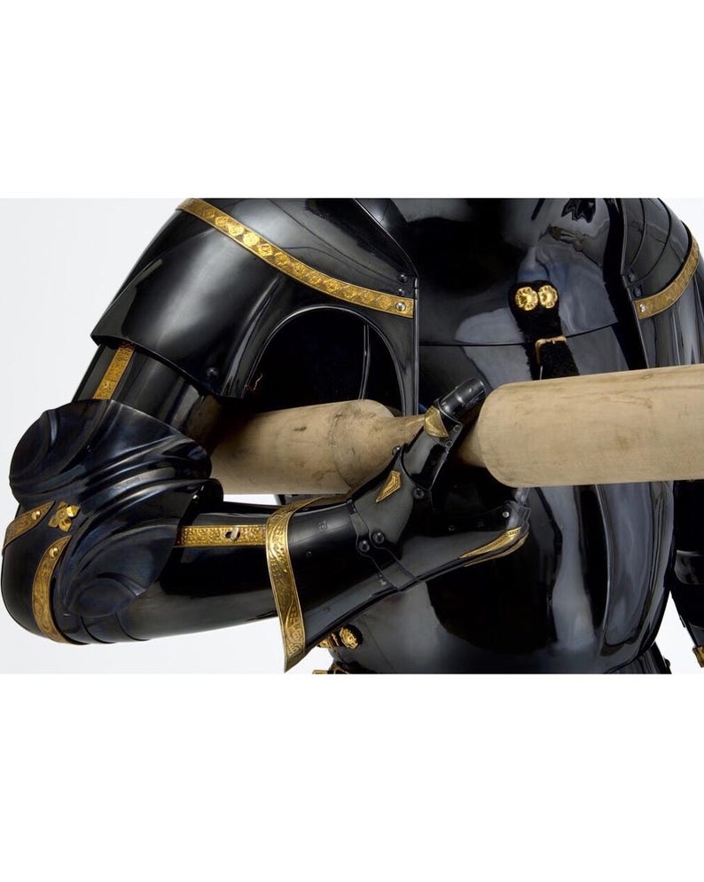 May include: Close-up of a black and gold medieval suit of armor. The armor features intricate gold detailing along the edges and joints. The gauntlets are holding a wooden object. The armor is highly polished and reflects the light.