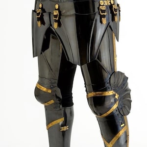 May include: Close-up of black metal armor with gold accents. The armor features articulated plates, decorative buckles, and knee protection. The design suggests a historical or fantasy theme, possibly for cosplay or display.