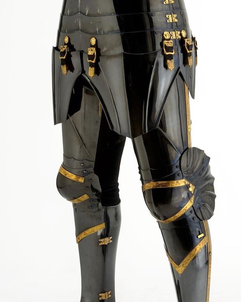 May include: Close-up of black metal armor with gold accents. The armor features articulated plates, decorative buckles, and knee protection. The design suggests a historical or fantasy theme, possibly for cosplay or display.