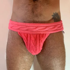 May include: A bright pink knitted jockstrap with a cable knit waistband and pouch. The jockstrap is made of a soft, textured yarn. The garment is designed for comfort and support.