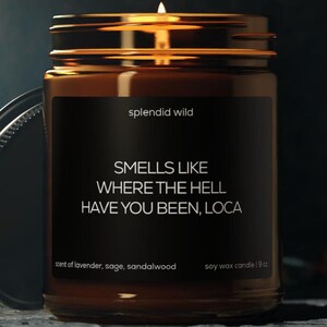 May include: A lit soy wax candle in a clear glass jar with a black label. The label reads "splendid wild" and "SMELLS LIKE WHERE THE HELL HAVE YOU BEEN, LOCA." The candle is scented with lavender, sage, and sandalwood, and is 19 oz.