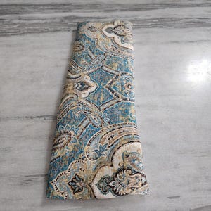 May include: Folded fabric with a detailed paisley pattern in shades of teal, beige, and brown. The fabric has a textured appearance, with intricate floral and curved designs. The overall aesthetic is vintage and decorative.
