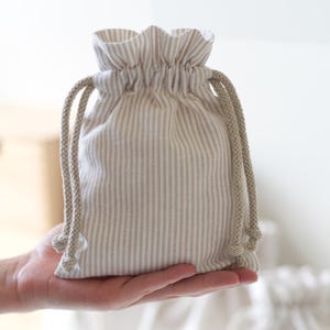 May include: A small drawstring pouch featuring a vertical beige and white striped pattern. The bag is constructed from fabric with a gathered top edge and includes two thick, braided beige rope cords on each side for closure and carrying purposes.