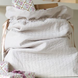 Könnte beinhalten: Eine große, beige gestreifte Stofftasche mit Kordelzug. Die Tasche ist mit Geschenken gefüllt und mit einem hellbraunen Band gebunden. Mehrere verpackte Geschenke mit floralen Mustern sind sichtbar.