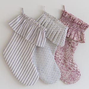 May include: Three decorative Christmas stockings with ruffled tops hang against a white wall. The stockings feature different patterns: blue and white stripes, a small blue floral print, and a burgundy floral print. Each stocking has a loop for hanging.