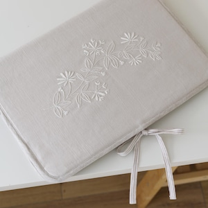 May include: A rectangular, light beige fabric cover featuring delicate white floral embroidery across the center. The cover has piped edges and includes two thin, striped fabric ties at the bottom corner for securing the item in a closed position.