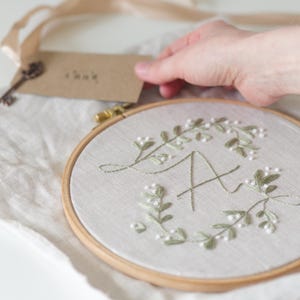 Pode incluir: Arte de bordado em bastidor com a letra "A" rodeada por uma coroa de folhas verdes e detalhes florais brancos. O bordado está num bastidor de madeira. Uma etiqueta com texto é segurada perto.