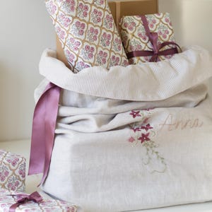 May include: A large, light-colored linen gift bag with a drawstring closure, holding wrapped gifts. The bag is embroidered with flowers and the name "Anna." A long, purple ribbon hangs from the bag. The gifts are wrapped in floral patterned paper.