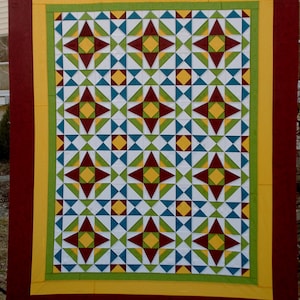 May include: A colorful quilt with a geometric star pattern. The quilt features a white background with repeating star designs in shades of red, yellow, green, and teal. The quilt is framed with a red border, yellow and green accents.