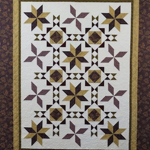 May include: A handcrafted quilt with a repeating star pattern in shades of brown, gold, and purple on a white background. The quilt has a floral border and a quilted texture, showcasing a traditional design.