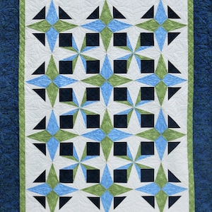 May include: A quilted textile featuring a repeating star pattern in shades of blue, green, and black on a white background. The quilt has a green border, and a dark blue border. The overall design is geometric and symmetrical.