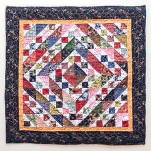 May include: A square patchwork quilt featuring a central diamond design in a variety of colours, including red, blue, green, and pink. It has a gold border and a dark blue patterned edge. Likely used as a decorative wall hanging.