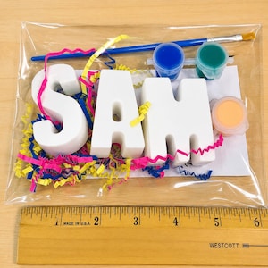 May include: A craft kit in a clear plastic bag containing three white letters spelling SAM, a blue paintbrush, and three small paint pots in blue, green, and peach. A wooden ruler shows the letters are approximately 5 inches wide in total.