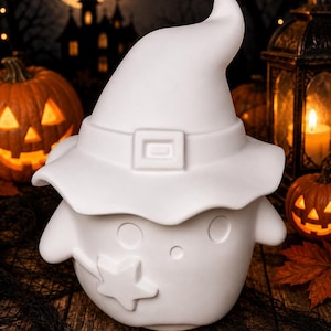 May include: A white ceramic figurine shaped like a ghost wearing a wide-brimmed witch hat. The ghost features two circular eyes, a small circular mouth, and a star-shaped detail on its side. The surface is matte white and unpainted.