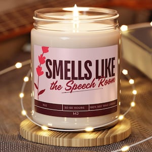 May include: A clear glass jar candle with a lit wick and a light pink label. The label features a red floral graphic and the text "SMELLS LIKE the Speech Room" in dark brown lettering, along with "9oz", "50-60 HOURS", and "100% SOY WAX CANDLE".