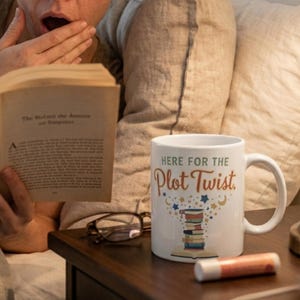 May include: A woman in bed reacts with surprise while reading a book. A white mug with the text "HERE FOR THE Plot Twist" and a stack of books is on the nightstand. The scene is lit by a bedside lamp.
