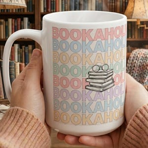 May include: A white ceramic mug with the word "BOOKAHOL" in pastel colors repeated vertically. The mug features a stack of books and glasses graphic. The mug is held in front of a bookshelf.