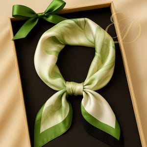 Op de afbeelding: Een zijden sjaal met een groen en wit bloemmotief, gepresenteerd in een geschenkdoos met een groene strik. De sjaal heeft een groene rand met een zwart accent. De sjaal is opgevouwen en geknoopt.