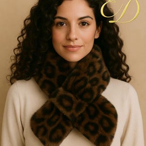 Puede incluir: Bufanda con estampado de leopardo marrón y negro. La bufanda está hecha de un material suave y afelpado y está envuelta alrededor del cuello. El fondo es de color beige liso.