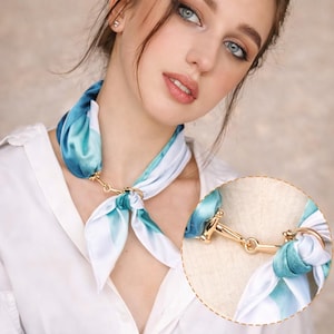 May include: A white and blue silk scarf with a gold-colored clasp. The scarf features a watercolor-like design in shades of blue and white. The clasp is a decorative element that connects the two ends of the scarf.