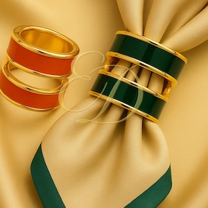 Könnte beinhalten: Zwei Sets Serviettenringe. Ein Set ist rot mit goldenem Rand, das andere ist grün mit goldenem Rand. Die Ringe befinden sich auf einer goldenen und grünen Serviette. Die Serviettenringe sind ein dekoratives Element für einen Esstisch.
