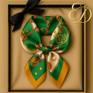 Peut inclure: Un foulard en soie vert et or avec un motif de chaîne et de volutes, présenté dans un coffret cadeau avec un nœud noir. Le foulard a une bordure crème et des accents dorés. Les initiales "E & D" sont en or en haut à droite.