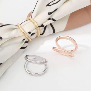 May include: Gold, rose gold, and silver scarf rings. The gold ring is holding a white scarf with black abstract designs. The rose gold and silver rings are displayed on a white surface. These rings are a stylish accessory.