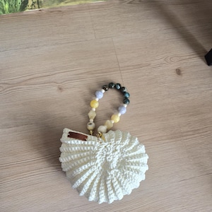 May include: A handmade, off-white crochet handbag with a seashell design. The bag features a beaded handle with a mix of colorful beads. The bag is sitting on a wooden surface.