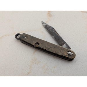 May include: A folding pocket knife with a dark, weathered metal handle featuring a textured, distressed finish and a small circular loop at one end. The blade is partially extended, showing a dark, oxidized surface with visible signs of wear.