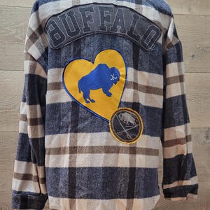 May include: Plaid flannel shirt in blue, brown, and white. Features a large "BUFFALO" patch across the shoulders and a heart-shaped patch with a blue bison. A smaller patch with crossed swords and a bison is also visible.