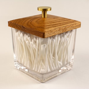 Op de afbeelding: Een heldere glazen pot gevuld met witte wattenstaafjes, bekroond met een vierkant houten deksel en een goudkleurige knop. De pot heeft een vierkante basis en is gevuld met talrijke witte wattenstaafjes.