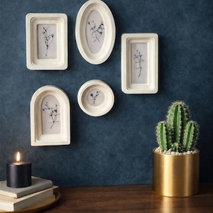 May include: A collection of five white picture frames of various shapes, each containing a delicate botanical print with blue accents, mounted on a dark blue wall. A gold planter with a cactus and a lit black candle on a wooden surface complete the decor.