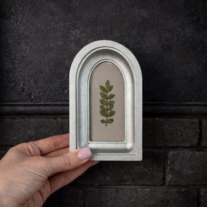 Puede incluir: Un marco de fotos plateado en forma de arco con un diseño de hojas verdes prensadas. El marco tiene un acabado texturizado y un fondo de color neutro. El marco está sostenido por una persona.