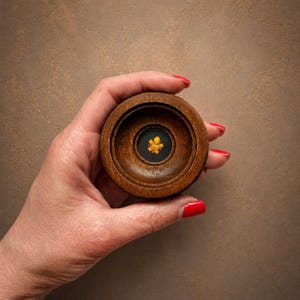 May include: A circular dark brown wooden container with a smooth, polished finish. The centre of the lid features a small, recessed circular inlay of black material, which holds a single, vibrant yellow pressed flower petal at its very centre.