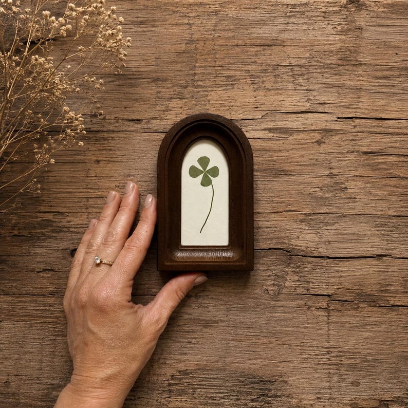 May include: A dark brown wooden arched frame containing a pressed green four-leaf clover on a cream-colored background. The frame features a smooth, rounded top edge and a rectangular base, showcasing a natural wood grain texture throughout the piece.