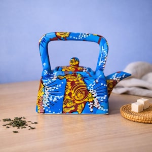 Puede incluir: Una tetera de cerámica azul con un vibrante estampado floral en amarillo y rojo. La tetera tiene un asa y un pico curvos, con un pequeño pomo en la tapa. El diseño incluye detalles en blanco. La tetera está sobre una superficie de madera.