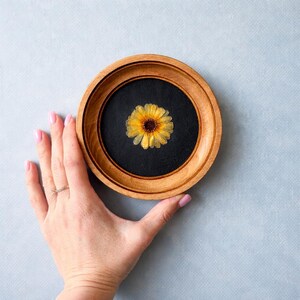 K&ouml;nnte beinhalten: Ein runder Holzrahmen mit schwarzem Innenraum h&auml;lt eine getrocknete gelbe Blume. Die Blume hat eine dunkle Mitte und gelbe Bl&uuml;tenbl&auml;tter. Der Rahmen wird von einer Hand mit rosa Nagellack vor einem hellblauen Hintergrund gehalten.