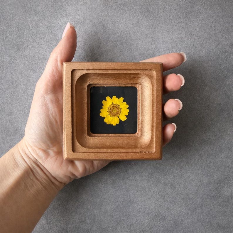 May include: A square wooden frame with a recessed center holding a yellow flower against a black background. The frame has a warm, natural wood tone. The flower is centered and in full bloom, with detailed petals.