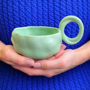 Puede incluir: Una taza de cerámica verde claro con un asa redonda. La taza se sostiene con dos manos sobre un fondo azul. La taza tiene una forma orgánica única y un acabado liso y mate.